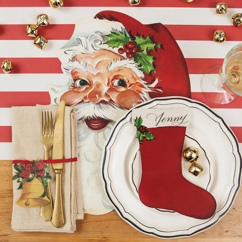 Christmas-themed table setting with Santa Claus face, red stocking table accent on a plate, and gold cutlery on a striped tablecloth.