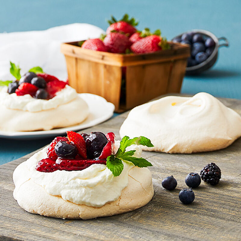 Three meringue pavais topped with whipped cream and mixed berries, arranged on a rustic wooden surface with a basket of strawberries and blueberries in the background.