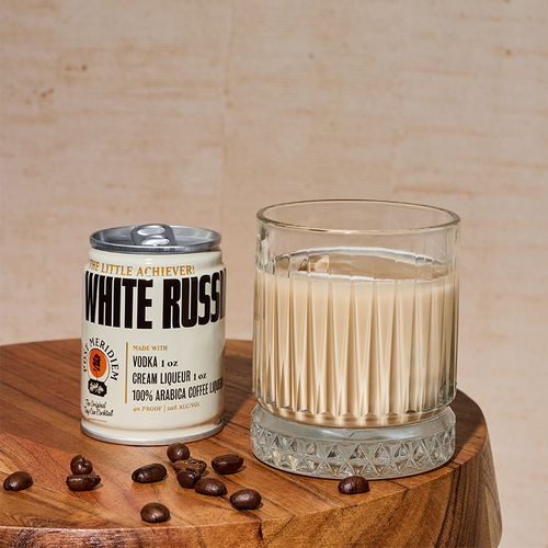 A can of White Russian sits beside a clear glass filled with the pale beige drink, set on a wooden surface with coffee beans scattered around. The background is a warm beige textured wall.
