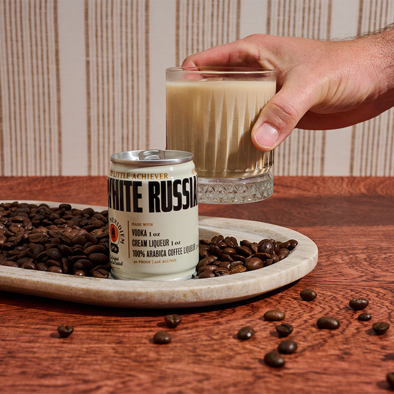 A hand holds a glass of creamy beige liquid behind a can labeled White Russian, set on a marble tray with coffee beans on a wooden table.