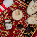 A styled flat-lay featuring a can of White Russian alcoholic cocktail, a glass with a creamy drink and ice cube, gold-framed sunglasses, and cream-colored sneakers on a red and black patterned rug.
