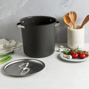 A large black stock pot on a kitchen counter next to its lid, a plate of ingredients, and a container with wooden utensils.