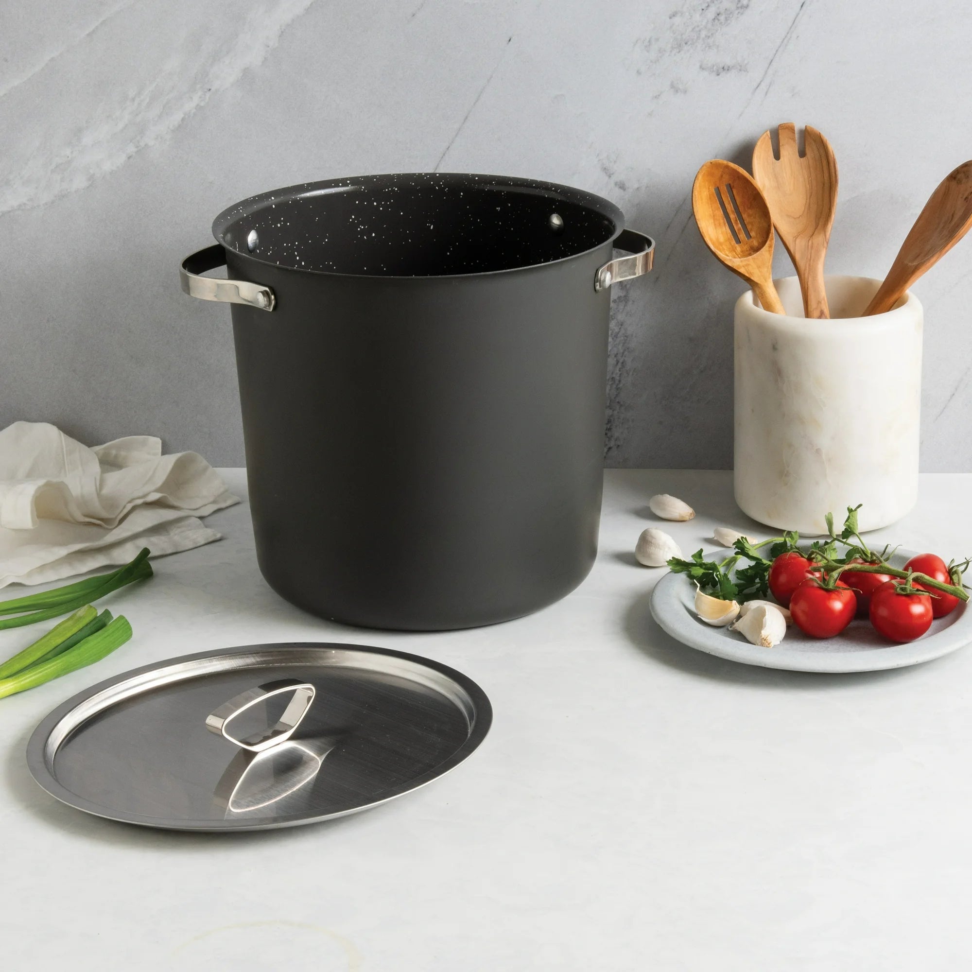 A large black stock pot on a kitchen counter next to its lid, a plate of ingredients, and a container with wooden utensils.