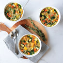 Three bowls of chunky soup, two on a white surface and the third on a round serving board with napkin, spoons, and sprig of rosemary.
