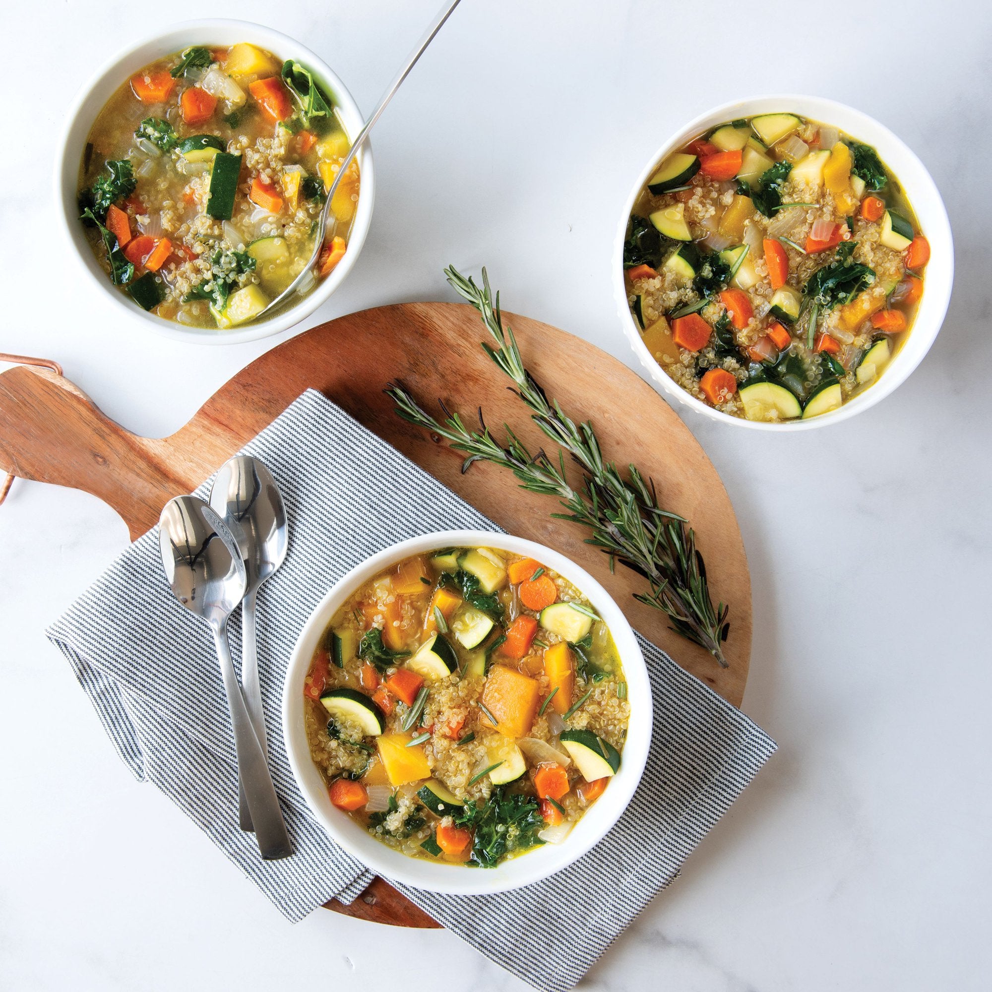Three bowls of chunky soup, two on a white surface and the third on a round serving board with napkin, spoons, and sprig of rosemary.