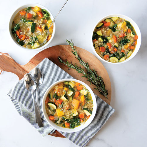 Three bowls of chunky soup, two on a white surface and the third on a round serving board with napkin, spoons, and sprig of rosemary.
