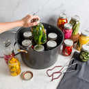A hand is shown placing a glass jar into a black canning pot filled with other preserved vegetables. Several glass mason jars with lids are arranged around a white marble countertop, alongside jar lids, canning tongs, and a dark cloth.