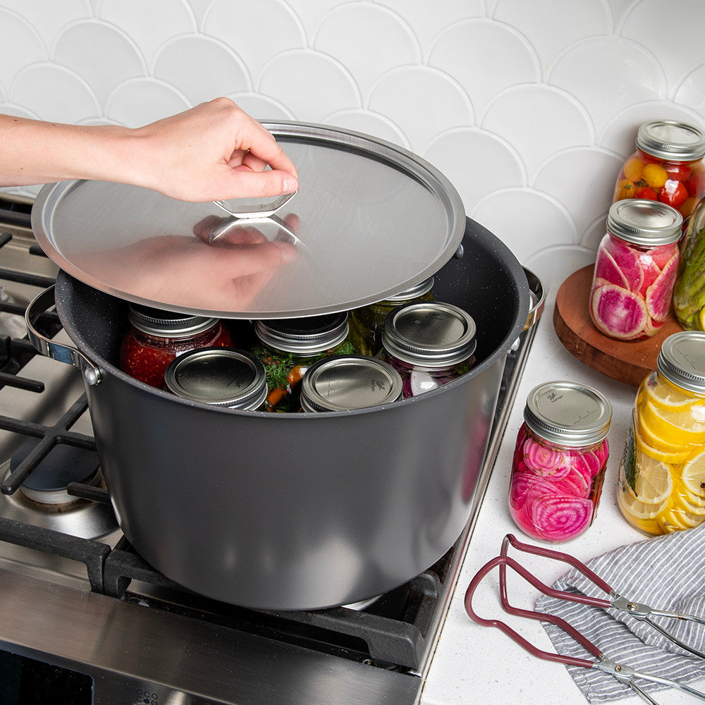 A large dark gray stockpot on a gas stove with a glass lid being lifted by a hand, revealing several glass canning jars inside. A white scalloped tile backsplash and a countertop with additional jars of pickled vegetables are visible in the background.