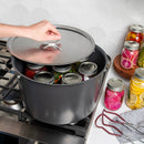 A large dark gray stockpot on a gas stove with a glass lid being lifted by a hand, revealing several glass canning jars inside. A white scalloped tile backsplash and a countertop with additional jars of pickled vegetables are visible in the background.