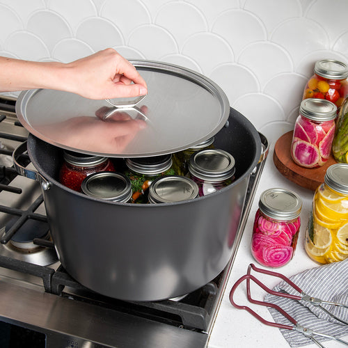 A large dark gray stockpot on a gas stove with a glass lid being lifted by a hand, revealing several glass canning jars inside. A white scalloped tile backsplash and a countertop with additional jars of pickled vegetables are visible in the background.
