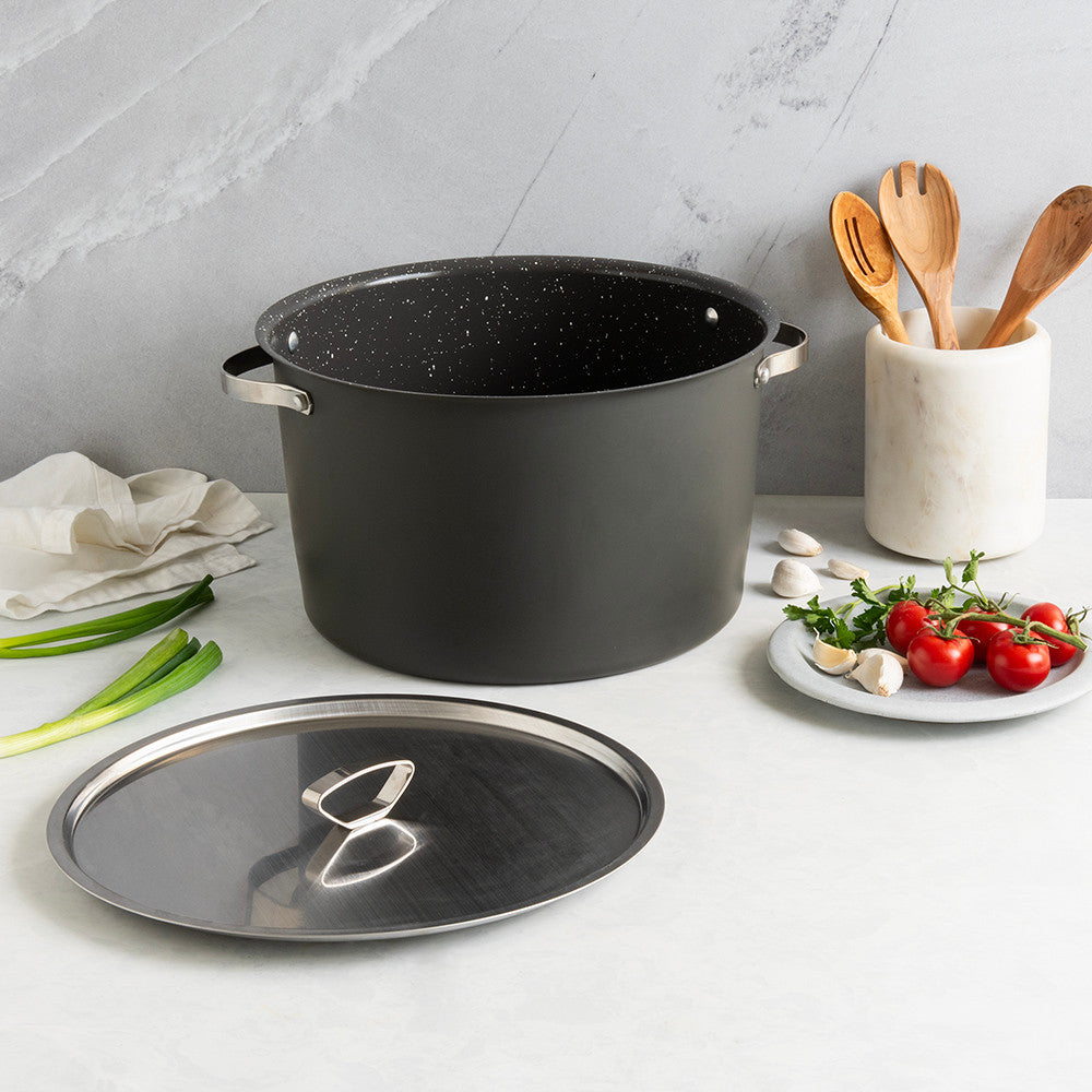 Black stock pot with lid on a kitchen counter beside a plate of ingredients and a container of wooden utensils.