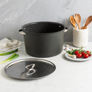 Black stock pot with lid on a kitchen counter beside a plate of ingredients and a container of wooden utensils.