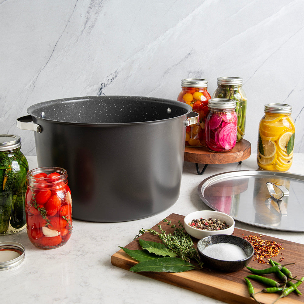 A large black enamel stockpot with silver handles sits on a white marble countertop, surrounded by glass jars of pickled vegetables, a metal lid, and cooking ingredients on a wooden cutting board.