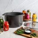 A large black enamel stockpot with silver handles sits on a white marble countertop, surrounded by glass jars of pickled vegetables, a metal lid, and cooking ingredients on a wooden cutting board.