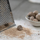 A grated nutmeg next to a grater and a bowl of nutmegs on a white surface.