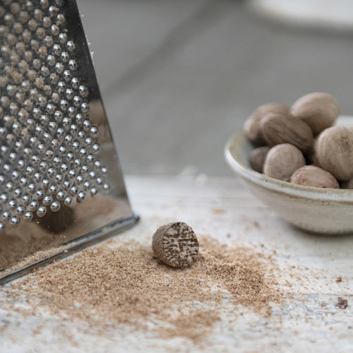 A grated nutmeg next to a grater and a bowl of nutmegs on a white surface.