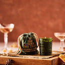 A small candle in a green votive beside a drawstring bag with 'WINE UNDER THE TREE' stitched onto it, both on a wooden block with champagne glasses in the background.