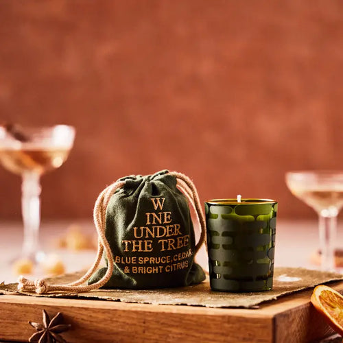 A small candle in a green votive beside a drawstring bag with 'WINE UNDER THE TREE' stitched onto it, both on a wooden block with champagne glasses in the background.