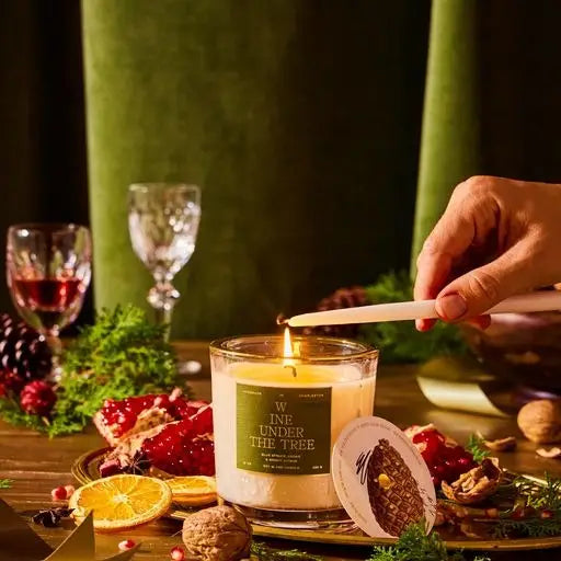 A person lighting a candle with a wine glass and various festive food items in the background, including a bowl of fruit and a glass of red wine.