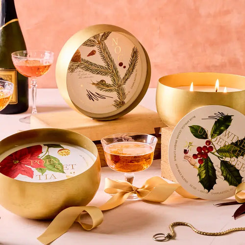 A person lighting a candle with a wine glass and various festive food items in the background, including a bowl of fruit and a glass of red wine.