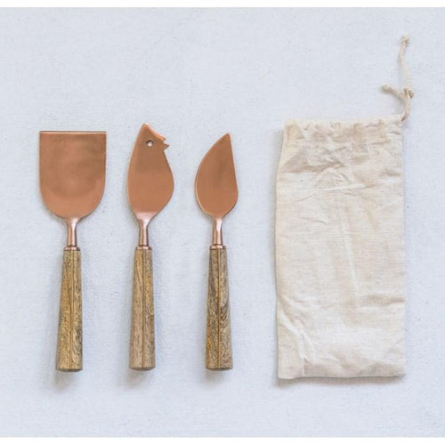 Three copper-colored cheese knives with wooden handles next to a drawstring bag on a white surface.