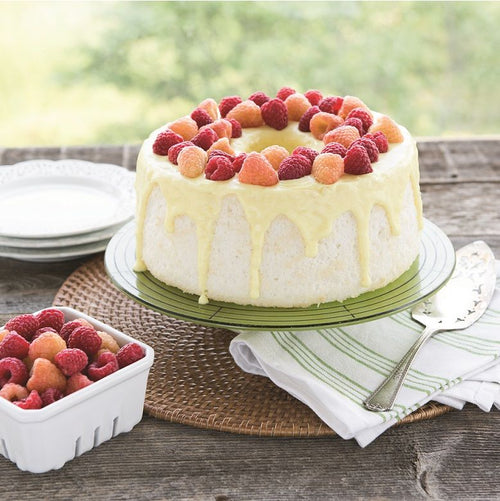 An angel food cake topped with icing and raspberries on a raised cake plate with a bowl of raspberries, plates, and cake server nearby on an outdoor wooden table.