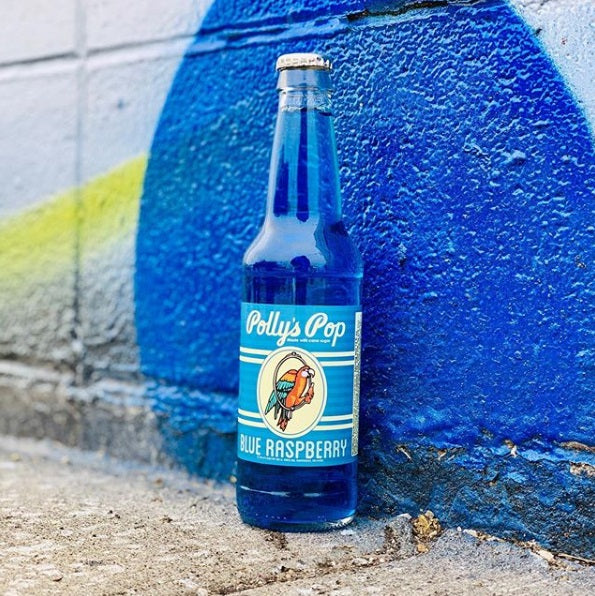 A bottle of blue raspberry Polly's Pop against a blue and white brick background.