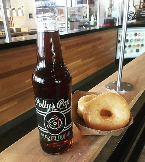 A bottle of glazed donut flavored Polly's Pop on a wooden surface next to a glazed donut.