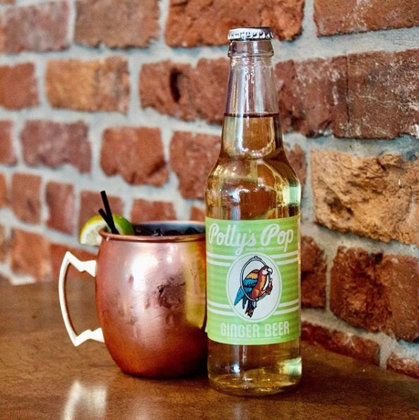 A bottle of ginger beer flavored Polly's Pop against a brick background next to a copper cup.