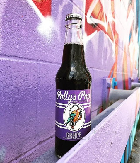 A bottle of grape Polly's Pop against a purple brick wall.