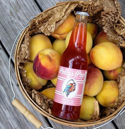 A bottle of peach Polly's Pop in a basket of peaches on a wood surface.