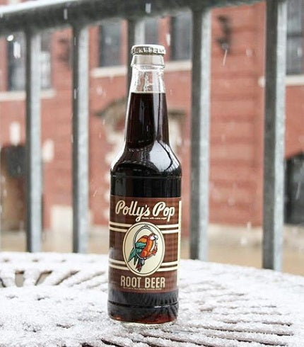 A bottle of root beer Polly's Pop on a snowy table with a blurred barred and brick background.
