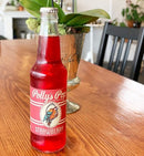 A bottle of strawberry Polly's Pop against on a wooden table with a chair and vase of plants in the background.