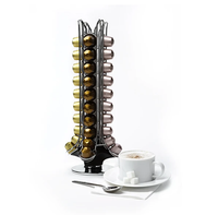 A silver-colored espresso carousel with multiple tiers, each holding gold-colored Nespresso capsules. The carousel is placed next to a cup of coffee and a saucer with dessert on it.