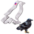A silver-colored cookie cutter in the shape of a crow, next to an example of a black crow cookie.