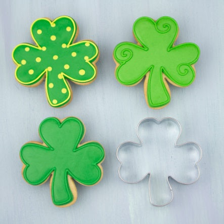 Three cookies decorated to look like clovers and a stainless steel shamrock cookie cutter on a gray background.