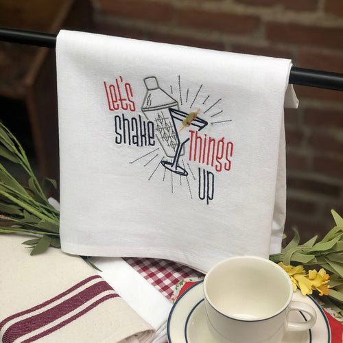 A white cotton tea towel with a colorful embroidered text design that reads 'Let's Shake Things Up' displayed on a table with a cup of tea and other kitchen towels in the background.