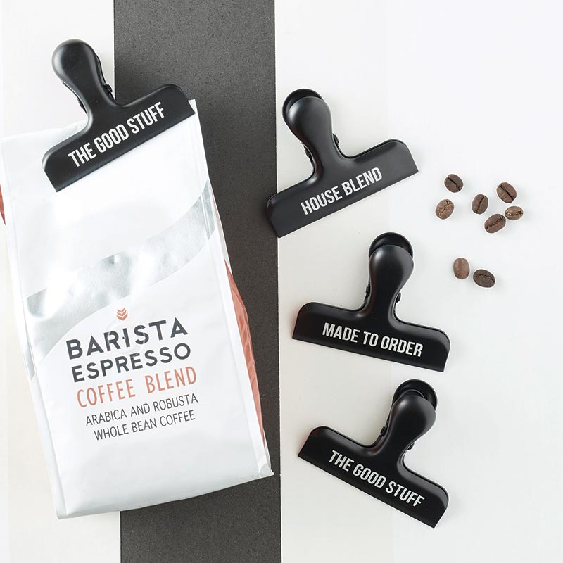 Several stainless steel black bag clips with a bag of barista espresso coffee blend and some coffee beans on a black and white background.