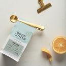 A coffee spoon doubling as a clip on a bag of Good Citizen coffee with an orange half and a bag clip on a neutral surface.