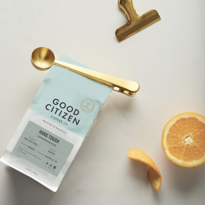 A coffee spoon doubling as a clip on a bag of Good Citizen coffee with an orange half and a bag clip on a neutral surface.