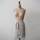 A cotton half apron with stripes displayed on a mannequin. The apron is primarily cream colored with black stripes and has a natural waistband.