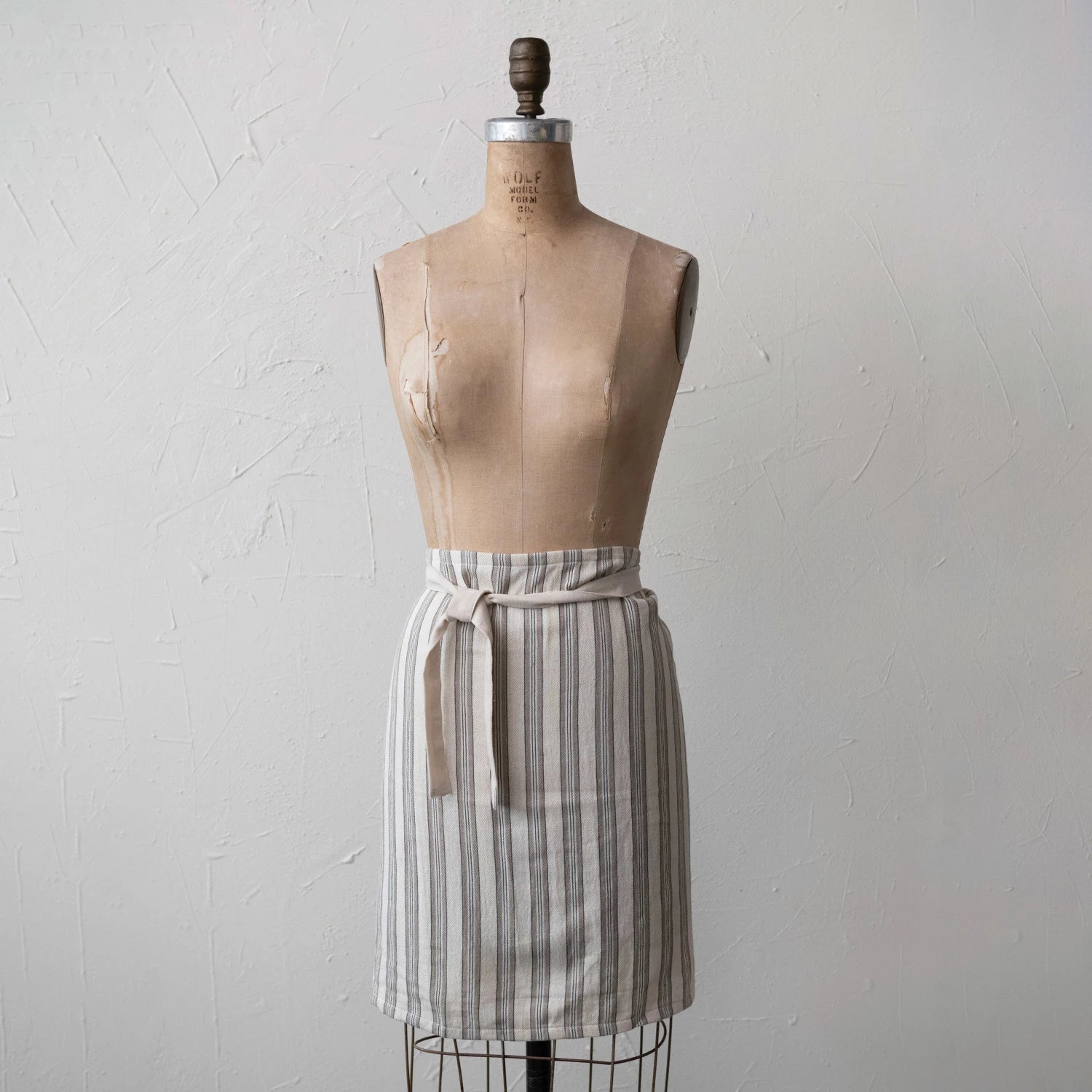 A cotton half apron with stripes displayed on a mannequin. The apron is primarily cream colored with black stripes and has a natural waistband.