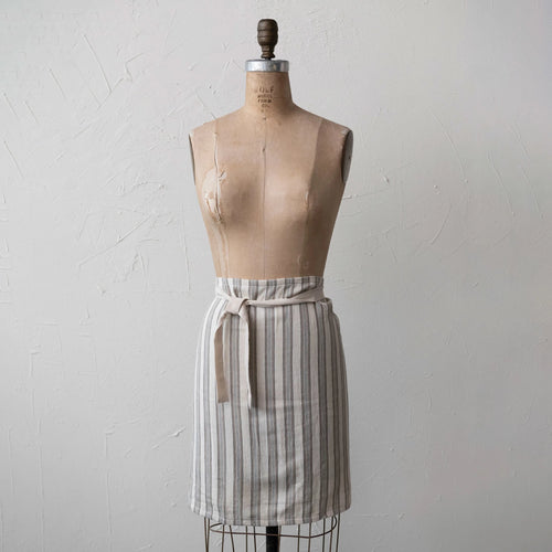 A cotton half apron with stripes displayed on a mannequin. The apron is primarily cream colored with black stripes and has a natural waistband.