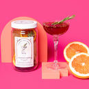 A jar of cocktail mix ingredients next to a finished cocktail and orange slices with a pink backdrop.