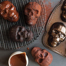 A skull cakelet pan next to a cooling rack with skull-shaped cakes and a pourer filled with chocolate sauce, all on a gray surface