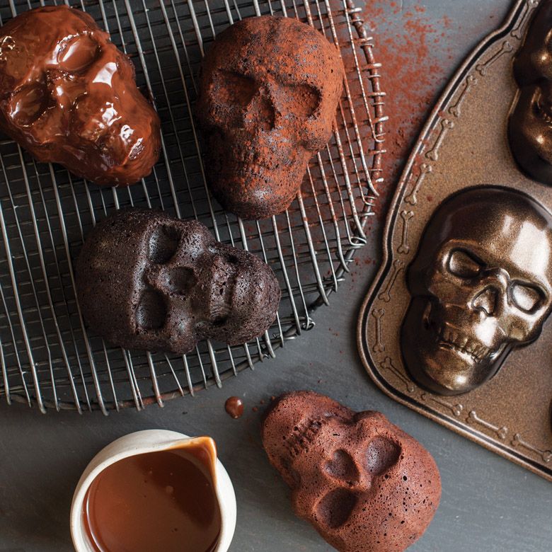 A skull cakelet pan next to a cooling rack with skull-shaped cakes and a pourer filled with chocolate sauce, all on a gray surface