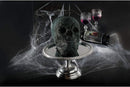 A dark cake mold of a skull on a silver display plate with glasses of white in the background with spiderwebs on a black surface.
