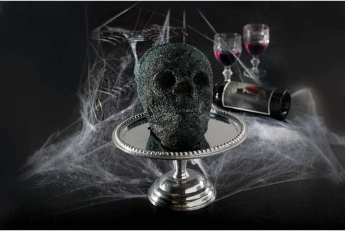 A dark cake mold of a skull on a silver display plate with glasses of white in the background with spiderwebs on a black surface.