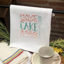 A white cotton tea towel with a colorful embroidered design that reads 'HAVE YOUR CAKE & EAT IT TOO' alongside a graphic of a fork, displayed in a kitchen setting with a cup and saucer.