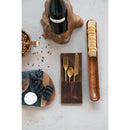 A leather cutlery sleeve with a golden fork, spoon, and knife inside next to wine and boards with food on them on a gray background.